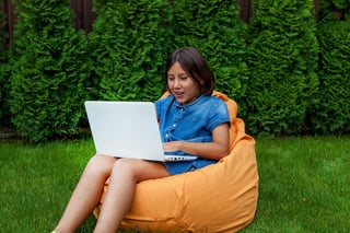 Girl Child Sits On A Pillow With An Open Laptop On The Green Grass In The Courtyard Of The