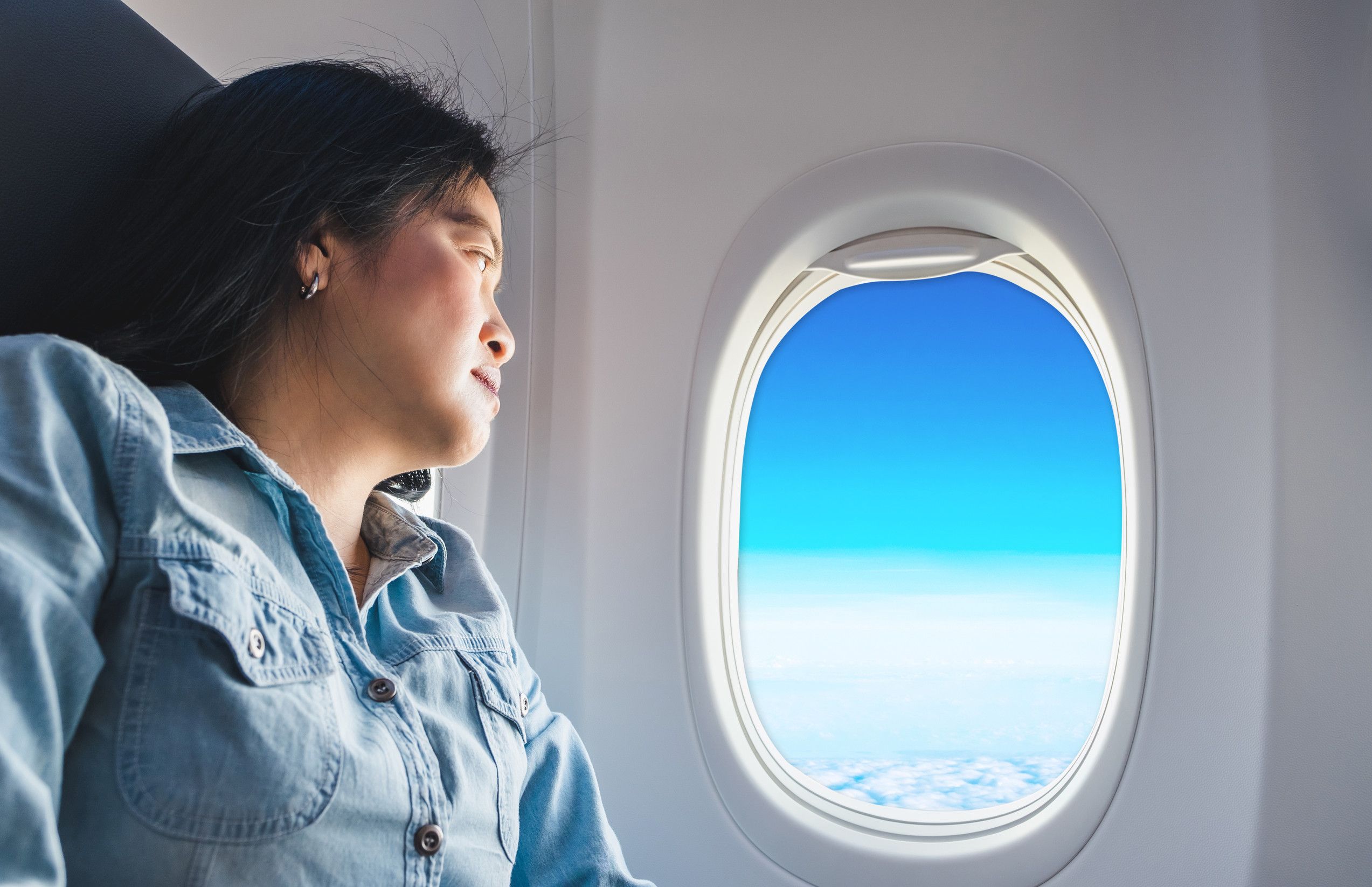 飛行機の窓側席に座るアジア人女性が窓の外を眺め、青空を眺める
